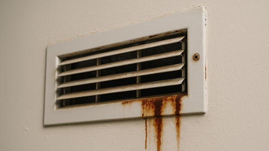 Old rusty air vent cover on a wall with brown corrosion stains and dust buildup.