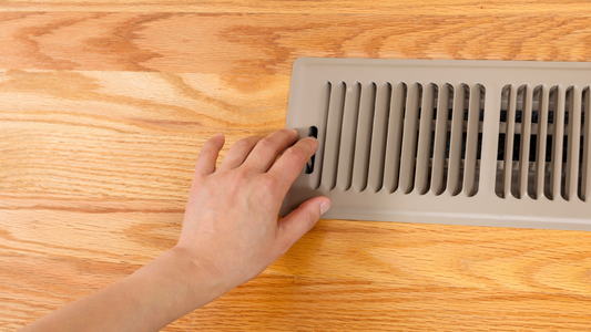 Hand adjusting brown floor register vent cover on wooden floor.